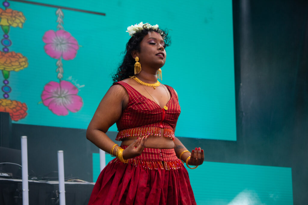 Optimal, a Tamil femme with deep skin tone and curly black hair, is wearing a red lehenga with beaded fringe and a crown of white flowers. 