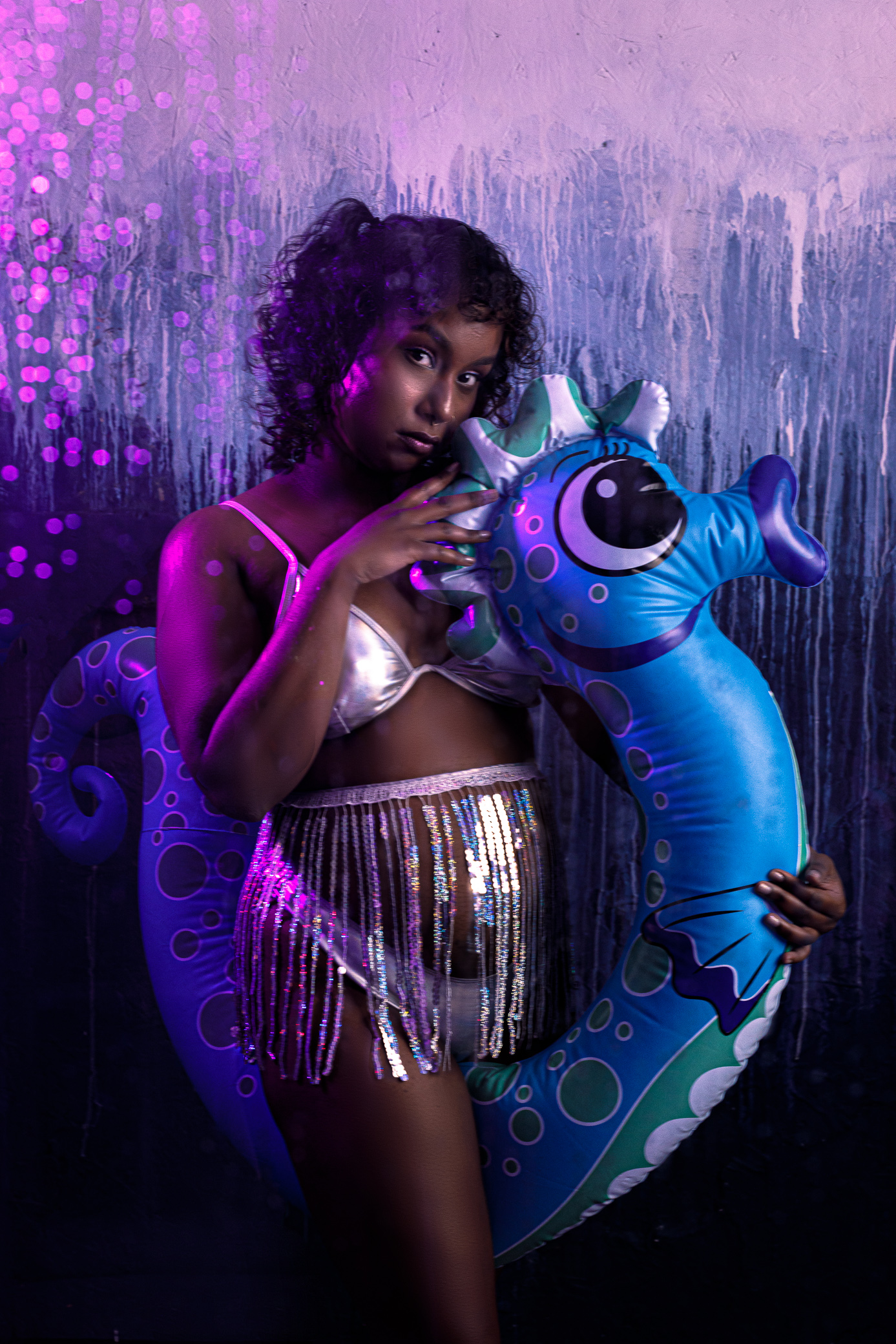 Optimal, a Tamil femme with a deep skin tone and curly black hair, us wearing a silver bikini and sequin fringe skirt. They are posing with a large inflatable blue seahorse, caressing the seahorse's face