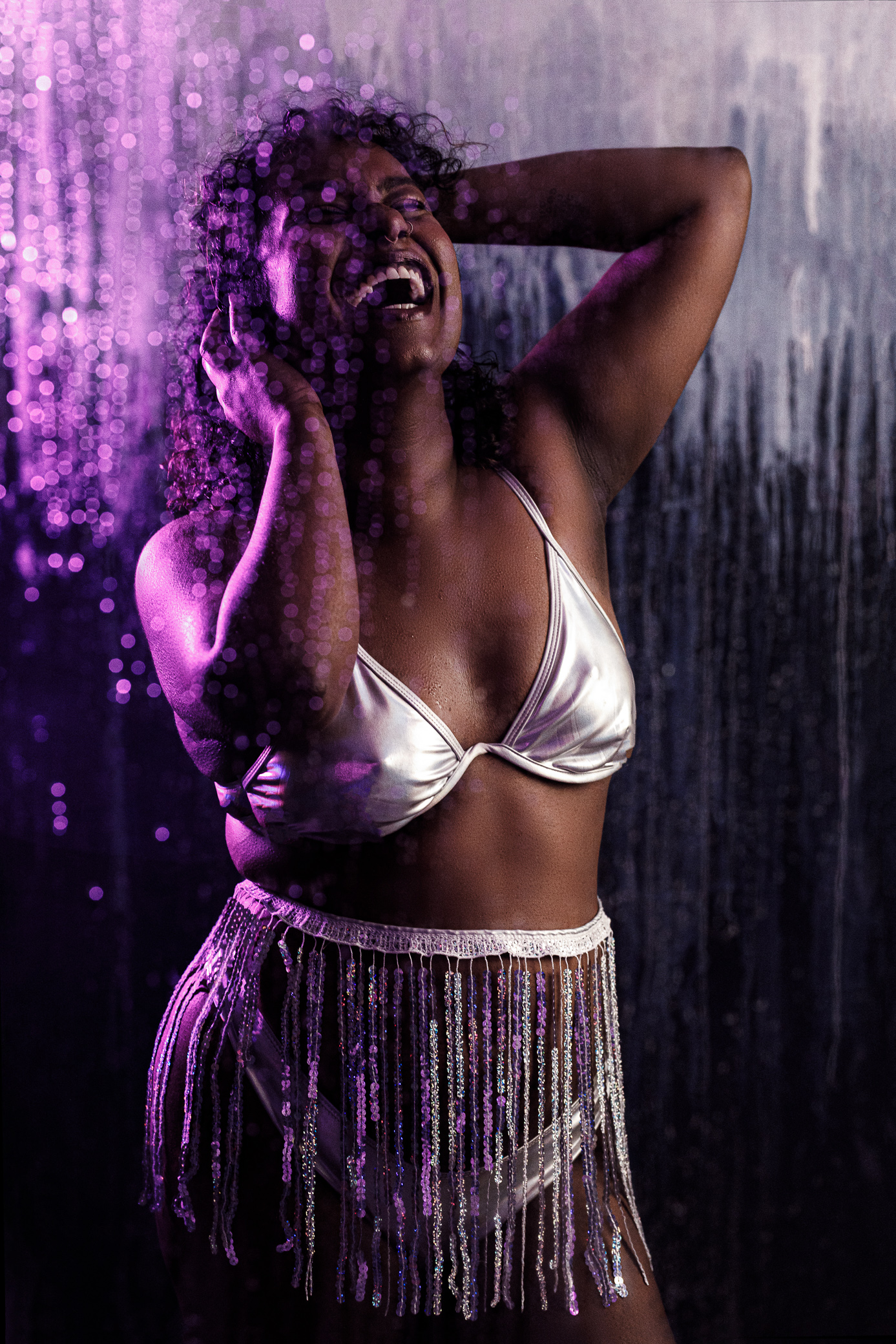 Optimal, a Tamil femme with a deep skin tone and curly black hair, us wearing a silver bikini and sequin fringe skirt. They are looking upwards with their left hand on their head and smiling